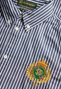 Marineblå og hvid stribet skjorte med knaplukning, der har en krave, hvide knapper og et broderet logo med krone og laurbærdetaljer.