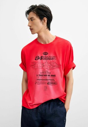 Giovane uomo che indossa una maglietta rossa con grafica, maniche arrotolate e pantaloni scuri, guarda verso sinistra su uno sfondo chiaro e uniforme.