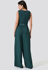 Grüner ärmelloser Jumpsuit mit tailliertem Oberteil, weitem Bein und einem Gürtel in der Taille. Verfügt über einen rückwärtigen Reißverschluss und einen sanften, leichten Stoff.