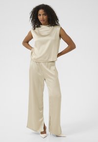 Haut sans manches en satin beige associé à un pantalon évasé, avec une coupe décontractée et des fentes latérales. Le modèle porte des talons pointus de couleur claire.