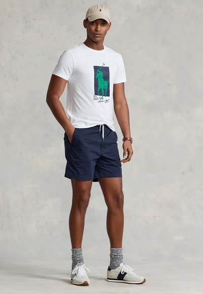 Vit bomullst-shirt med grön grafisk polo-spelare, marinblå shorts, ljusbeige keps, grå strumpor och vita sneakers med marinblå accenter.