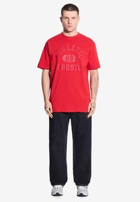 Czerwona bawełniana koszulka z grafiką "ATHLETIC 99 PROSTO" w kontrastowej czcionce. Noszona z luźnymi ciemnymi dżinsami i trampkami.