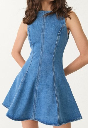 Kobieta ubrana w niebieską, dopasowaną sukienkę dżinsową bez rękawów z rozkloszowanym dołem na jednolitym jasnym tle.