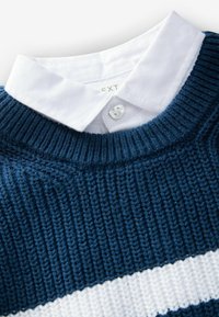 Blauer Strickpullover mit geripptem Muster, versehen mit einem weißen Streifen und einem weißen Kragenhemd darunter, das am Kragen zugeknöpft ist.
