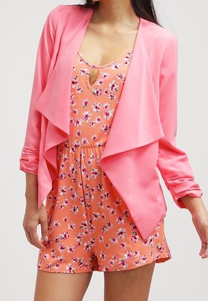 Blazer drapé léger rose sur une combinaison orange à motifs floraux avec un décolleté en forme de trou de serrure. Texture lisse avec une coupe décontractée.