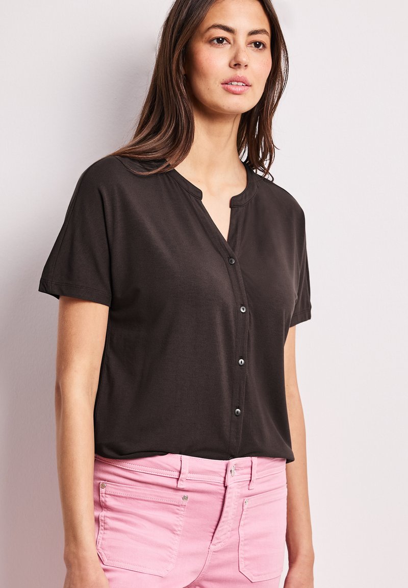 Kvinde med langt brunt hår iført en kortærmet sort skjorte med knapper og lyserøde bukser, stående mod en ensartet lys baggrund.