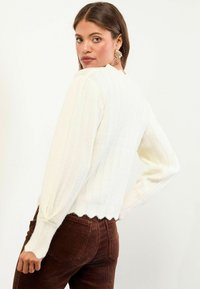 Barna hajú nő arany kagylóformájú fülbevalókkal, fehér bordázott pulóverben, hullámos szélekkel és barna kordbársony nadrágban, háttal áll.