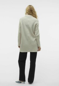 Maglione lungo grigio chiaro con taglio dritto, design a un solo bottone e spacchi laterali, abbinato a pantaloni neri e scarpe blu con punta.
