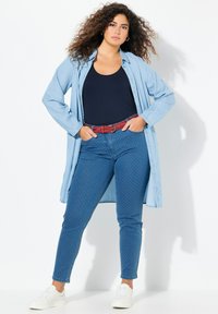 Camisa de mezclilla azul clara, camiseta sin mangas azul marino, jeans oscuros con un patrón de puntos blancos, cinturón tejido rojo y zapatillas blancas. Cabello rizado, pose de pie.