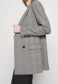 Blazer a cuadros gris con un corte holgado, que presenta líneas rojas y blancas, un botón negro en el centro y dos bolsillos frontales. Textura suave.
