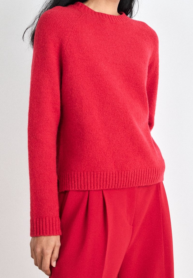 Pull rouge en maille avec un col rond, des poignets et un ourlet côtelés. Texture douce et coupe légèrement courte, associé à un pantalon rouge.