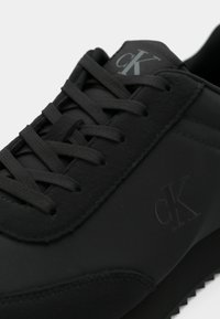 Prim-plan al unui sneaker negru Calvin Klein, evidențiind șireturile texturate, accentele din piele întoarsă și logo-urile „cK” embossate pe limbă și pe lateral.
