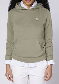 Olive green hoodie with a kangaroo pocket, featuring soft fabric and an embroidered logo. Worn over a blue-striped collared shirt.