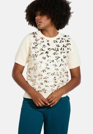 Maglione corto in crema con paillettes di varie tonalità, caratterizzato da una consistenza morbida e polsini a costine, abbinato a pantaloni teal.
