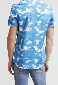 Mężczyzna w niebieskiej koszuli z krótkim rękawem z białymi abstrakcyjnymi wzorami, połączonej z jasnoniebieskimi dżinsami, pokazany od tyłu.