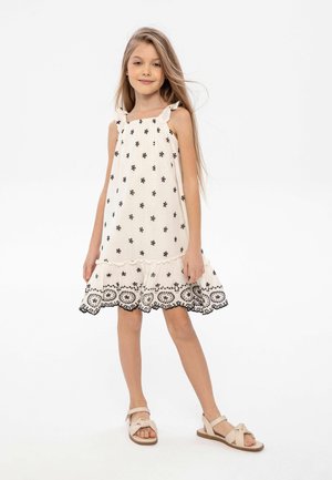 Robe sans manches crème avec des motifs floraux noirs, ourlet froncé et bord en dentelle décoratif. Associée à des sandales beige.