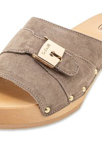 Bruna mockaskinnsandaler med ett guldspänne, synlig sömnad och träsoffa med dekorativa metallspikar.