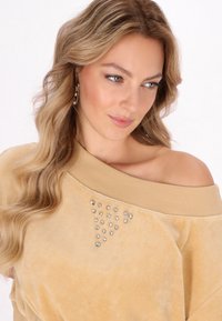 Beige, off-shoulder pulóver, puha tapintással, az elején kristály díszítésekkel, amelyek háromszög mintázatban vannak elhelyezve.