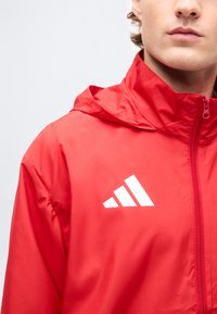 Νεαρό άτομο που φοράει κόκκινο αθλητικό μπουφάν με φερμουάρ και λευκό λογότυπο με τρεις ρίγες στο στήθος, πάνω σε απλό φόντο.