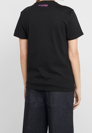 Zwart katoenen T-shirt met een ronde hals en korte mouwen. Op de achterkant zit een roze logodetail bij de hals. Gecombineerd met donkerblauwe spijkerbroek.