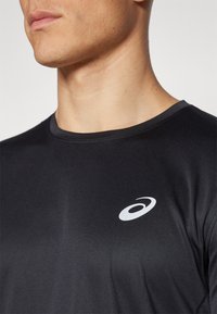 Läheltä kuva henkilöstä, joka käyttää mustaa urheilupaitaa, jossa on valkoinen Asics-logo rinnassa, näkyen kaula ja alaleuka.