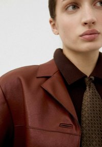 Blazer di pelle marrone con rever, abbinato a una maglietta a coste marrone scuro e una cravatta a fantasia nei toni del verde e marrone.