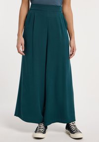 Pantaloni a gamba larga teal con pieghe, vita alta e tessuto morbido. Abbinati a sneaker alte nere.