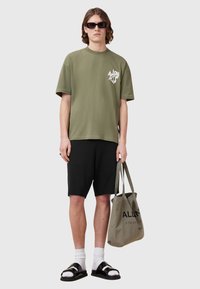 Olive green oversized t-shirt with logo, black shorts, white socks, black sandals, and a canvas tote bag with printed text. Neutral background.