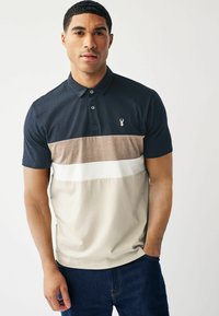 Polo majica z mornarsko modrim vrhom, bež in belimi vodoravnimi črtami. Ima manjši logo na prsih in klasičen ovratnik. Tkanina se zdi gladka.