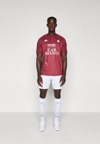 Bordeaux fotbaltrøye med korte ermer, hvite shorts og hvite sokker. Trøya har logoer og et subtilt mønster, med røde aksenter.