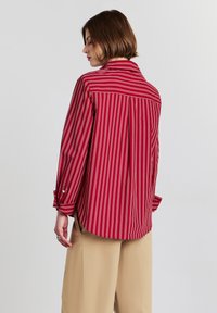 Camicetta a righe rosse con una vestibilità comoda, caratterizzata da maniche lunghe, colletto e orlo leggermente curvo. Abbinata a pantaloni beige.