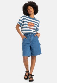 Rutet blå og hvit t-skjorte med korte ermer og grafikk av en oransje sol, sammen med blå denimshorts og svarte plattformsandaler.
