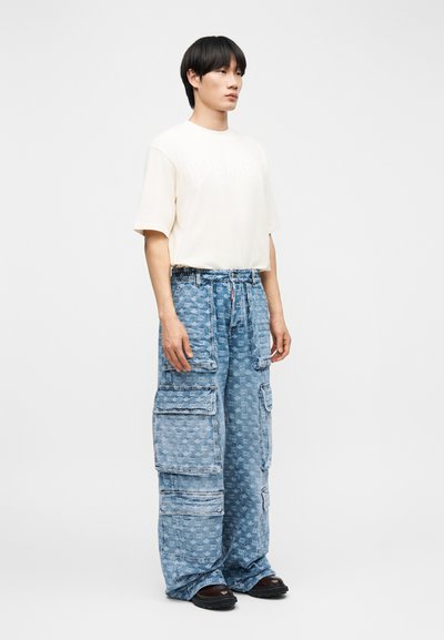 Jeune homme debout portant un t-shirt crème oversize et un jean cargo large bleu clair avec poches, ainsi que des chaussures noires, sur un fond uni.