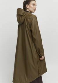 Trench de ploaie lung, de culoare verde oliv, realizat dintr-un material lucios și impermeabil. Dispune de o glugă, manșete ajustate și detalii cu nasturi pe față și pe laturi.