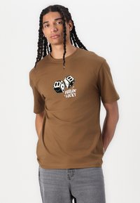 Brun bomulds T-shirt med grafik af to terninger med smilende ansigt og teksten "Feelin' Lucky" nedenunder. Korte ærmer, crew neck design.