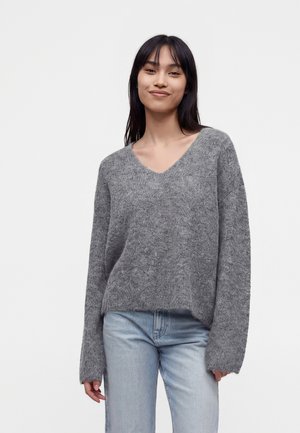 Jeune femme aux longs cheveux foncés portant un pull ample gris à col en V et un jean bleu clair, debout sur un fond uni.