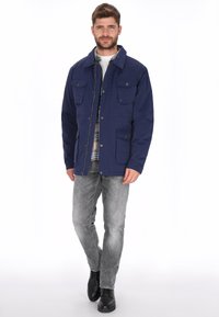 Chaqueta azul marino con cuello, múltiples bolsillos frontales, cierre de cremallera y material resistente, combinada con jeans grises y botas negras.