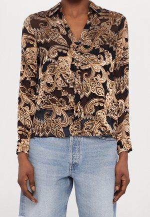 Blouse à manches longues à motif cachemire noir et beige avec col, portée avec un jean en denim bleu clair, mains détendues le long du corps.