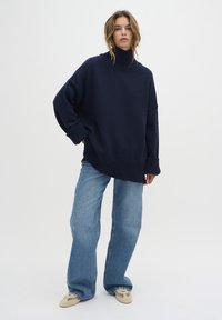 Marinblå oversized stickad tröja med hög ribbad krage och muddar, tillsammans med ljusblå vida jeans och beige skor.