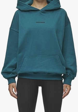 Personne portant un sweat à capuche couleur sarcelle avec une poche avant et des leggings noirs, debout, les mains détendues le long du corps.