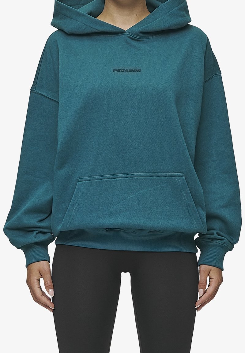 Personne portant un sweat à capuche couleur sarcelle avec une poche avant et des leggings noirs, debout, les mains détendues le long du corps.