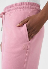 Roze joggingsbroek met een elastische tailleband en een koord, voorzien van een zijzak en een ronde logo patch op de bovenbeen. Zachte stoftextuur.