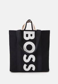 Svart tygväska med stora vita bokstäver "BOSS" på framsidan, med kontrasterande handtag i beige och svart.