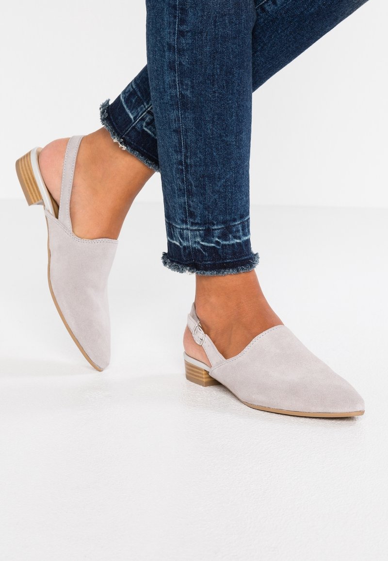 Pieds en ballerines slingback en daim gris clair avec petits talons en bois, associées à un jean skinny bleu foncé à ourlet effiloché sur un sol blanc.
