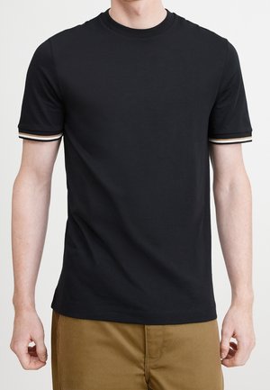 Mann trägt ein schlichtes schwarzes Kurzarm-T-Shirt mit gestreiften Bündchen an den Ärmeln und braune Hose, steht vor weißem Hintergrund.