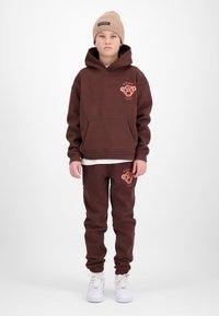 Kind draagt bruine hoodie en bijpassende joggingbroek met bedrukt berenlogo, beige gebreide muts en witte sneakers, staand op witte achtergrond.