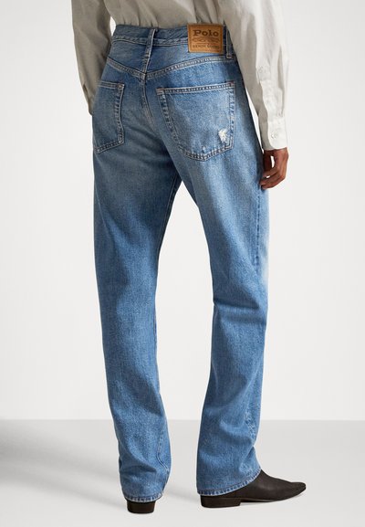 Tagantvaade inimesest, kes kannab helerohelisi teksaseid, millel on tagataskud ja Polo Denim Goods nahast silt, valget särki ja musti kingi.