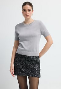 Lys grå kortærmet strikbluse med en struktureret, glitrende krave. Sammen med en sort pailletnederdel. Glat stof, tætsiddende design.
