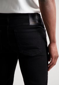 Vaqueros de mezclilla negra con corte ajustado, que presentan una etiqueta de cuero con la marca "DSTREZZED" en la parte de atrás, y detalles de costura sutiles en los bolsillos.