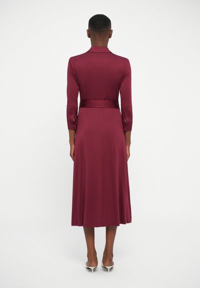 Bordo haljina napravljena od glatkog materijala, sa ovratnikom, dugim rukavima, strukom s remenom i lepršavom suknjom koja završava ispod koljena.